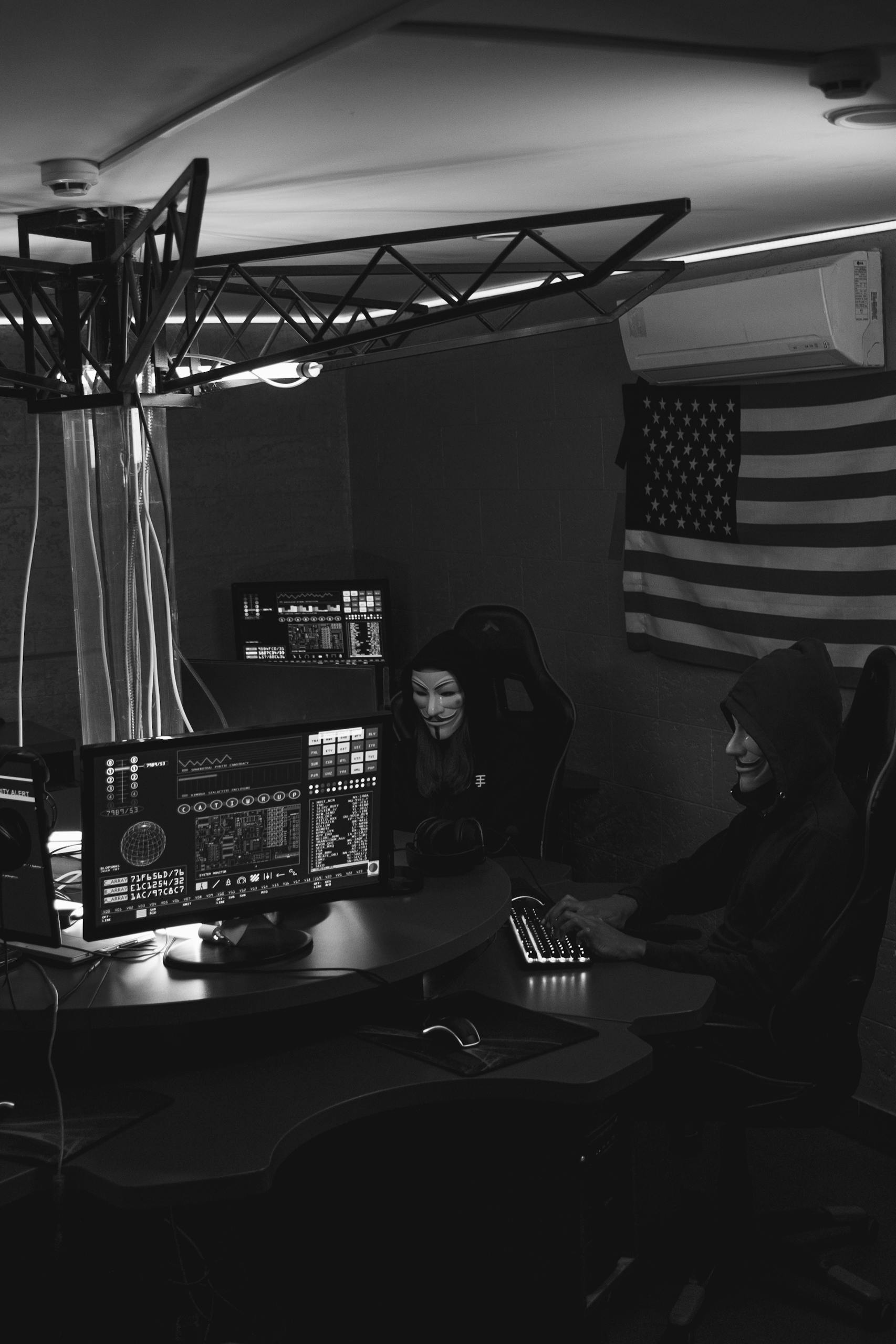 Two anonymous hackers in a dim room working on computers, symbolizing a cyber attack.